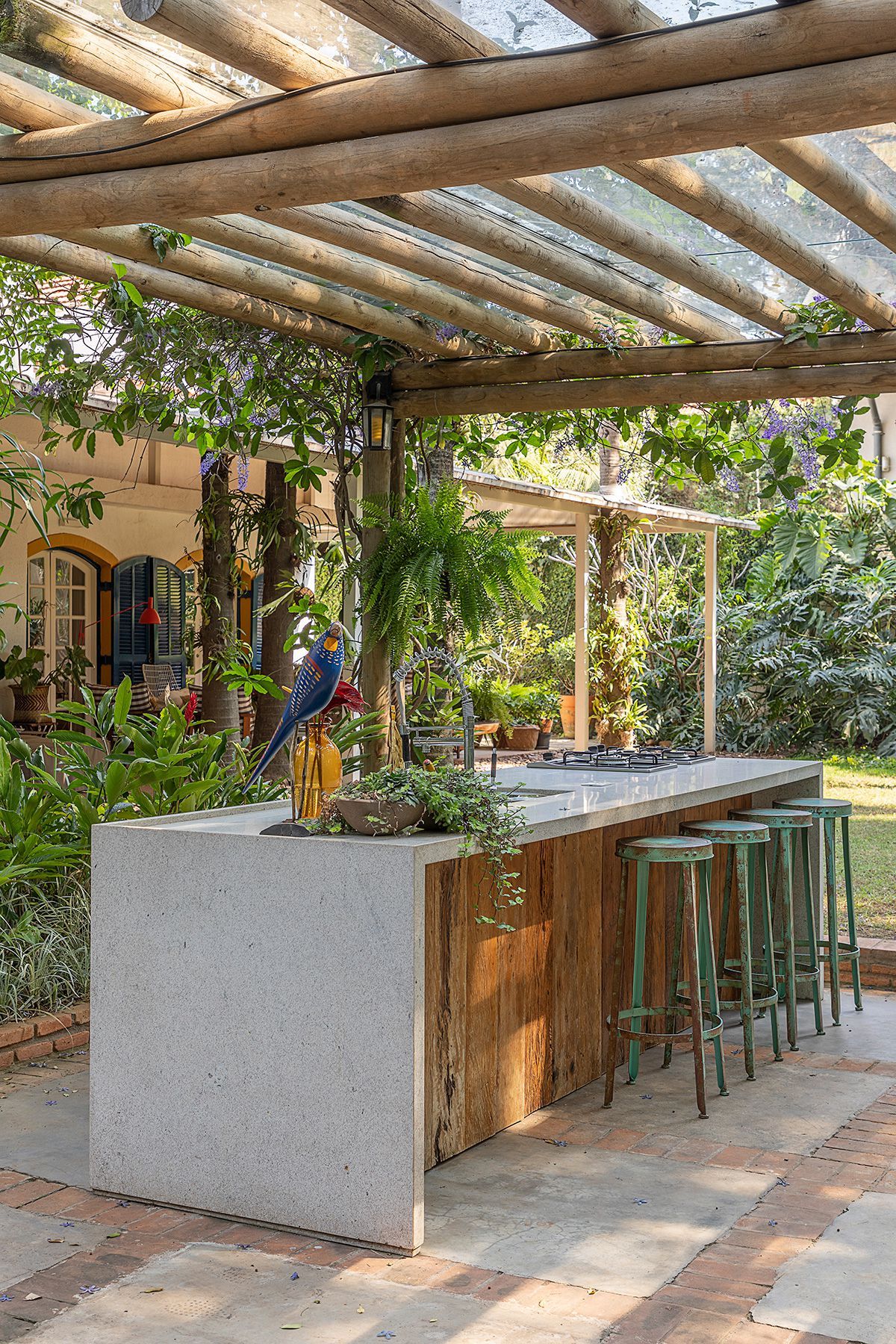 Espaço gourmet externo de inspiração tropical-colonial inserido em exuberante jardim sombreado, com bancada monolítica em pedra natural clara apoiada sobre base de madeira de demolição, equipada com cooktop a gás e ladeada por banquetas industriais em metal patinado verde-água. O pergolado em toras roliças de eucalipto com cobertura translúcida filtra a luz natural enquanto trepadeiras de tumbérgia-arbustiva com flores lilases se entrelaçam nas vigas, criando um microclima agradável junto a sama, estilo Tropical, Classico, elementos Bancada de cozinha gourmet externa em pedra natural clara (granito ou limestone) com base revestida em madeira de demolição, Banquetas altas industriais em metal patinado na cor verde-água com assento redondo, Pergolado em toras roliças de eucalipto tratado com cobertura translúcida em vidro ou policarbonato, Piso em tijolo cerâmico assentado em paginação espinha-de-peixe e cimento liso, Cooktop a gás embutido na bancada externa, Luminária tipo lanterna colonial em ferro forjado pendente no pergolado, Escultura decorativa de arara em cerâmica vitrificada policromada, Vasos em cerâmica rústica com plantas pendentes sobre a bancada, Janelas arqueadas com venezianas em madeira pintadas de azul na edificação ao fundo, Cabos elétricos embutidos nas toras do pergolado, plantas Samambaia-de-boston, Tumbérgia-arbustiva, Filodendro, Dinheiro-em-penca, Costela-de-adão, Helicônia, Moréia, Jacarandá-mimoso, Peperômia-pendente, ambiente acolhedor, sofisticado, relaxante, localização quintal, varanda, área medio, estruturas pergolado, churrasqueira, iluminacao, cores verde-escuro, verde-claro, cinza-claro, marrom-madeira, verde-água, azul-colonial, amarelo-ocre, terracota, piso pedra, pisantes, privacidade cerca_viva, água nenhum