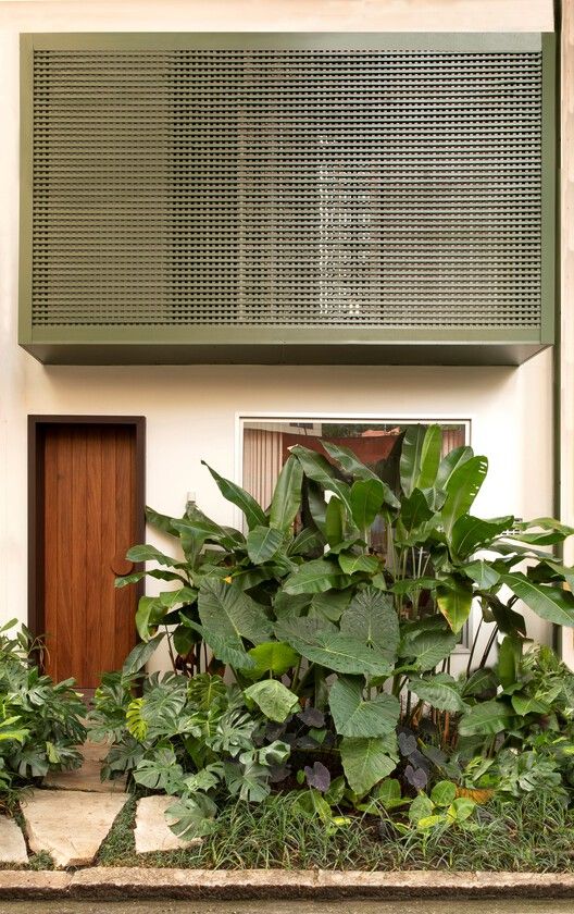 Jardim frontal de fachada residencial contemporânea com forte influência do paisagismo tropical brasileiro, onde um denso maciço de folhagens exuberantes — dominado por costelas-de-adão, alocásias, taiobas-roxas e bananeiras-ornamentais — cria uma cortina verde natural que confere privacidade e frescor à entrada da casa. A composição arquitetônica integra um elegante painel de cobogó metálico verde-oliva no pavimento superior com uma porta pivotante em madeira nobre, enquanto pisantes de pedra n, estilo Moderno, Tropical, elementos Porta pivotante em madeira maciça de tom escuro, Painel superior em cobogó metálico na cor verde-oliva, Parede rebocada e pintada em tom creme/off-white, Pisantes em pedra natural irregular (São Tomé ou similar), Canteiro frontal adensado com volumetria tropical, Janela com cortina em tecido translúcido atrás da vegetação, plantas Costela-de-adão, Taioba-ornamental, Helicônia, Bananeira-ornamental, Filodendro, Orelha-de-elefante, Grama-esmeralda, Taioba-roxa, ambiente moderno, sofisticado, acolhedor, localização fachada, entrada, jardim, área pequeno, estruturas nenhum, cores Verde-escuro, Verde-claro, Verde-oliva, Marrom-madeira, Creme, Bege, Roxo-escuro, piso grama, pisantes, pedra, privacidade muro_verde, água nenhum