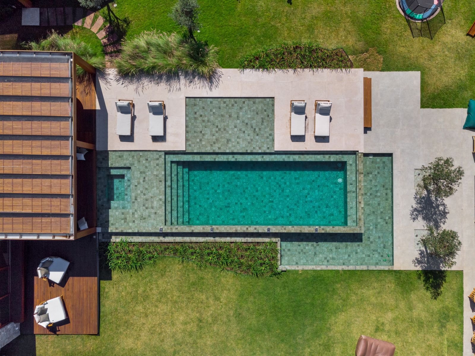 Projeto paisagístico contemporâneo de alto padrão com piscina de borda infinita revestida em pedra Hijau, integrando prainha de acesso gradual e área profunda em formato retangular, cercada por piso atérmico de porcelanato cimentício de grande formato. O jardim combina extensos gramados de Zoysia japonica com massivos tropicais de bambuzinhos, capim-do-texas e heliconias dispostos em canteiros lineares que suavizam a transição entre a área molhada e os espaços verdes. O deck em madeira cumaru co, estilo Moderno, Tropical, elementos Piscina retangular com borda infinita e degraus internos em pedra natural, Prainha/beach entry com revestimento em pedra Hijau (pedra Bali), Deck elevado em madeira cumaru com estrutura de pergolado, Quatro espreguiçadeiras brancas com almofadas sobre piso atérmico, Piso em porcelanato cimentício de grande formato no entorno da piscina, Canteiro linear elevado com vegetação tropical entre gramado e piscina, Mesa e cadeiras em área de estar no deck lateral, Cama de rede/trampoline circular no gramado superior direito, Escada caracol metálica no canto superior esquerdo, Iluminação embutida no paisagismo perimetral, plantas Grama-esmeralda, Palmeira-fênix, Helicônia, Moreia, Capim-do-texas, Agapanto, Formio, Clúsia, Oliveira, Bambuzinho-de-jardim, Liríope, Podocarpo, ambiente sofisticado, moderno, relaxante, localização piscina, jardim, quintal, área grande, estruturas pergolado, iluminacao, cores verde-esmeralda, turquesa, bege, branco, cinza, madeira-natural, verde-escuro, piso grama, deck, porcelanato, pedra, privacidade cerca_viva, água piscina