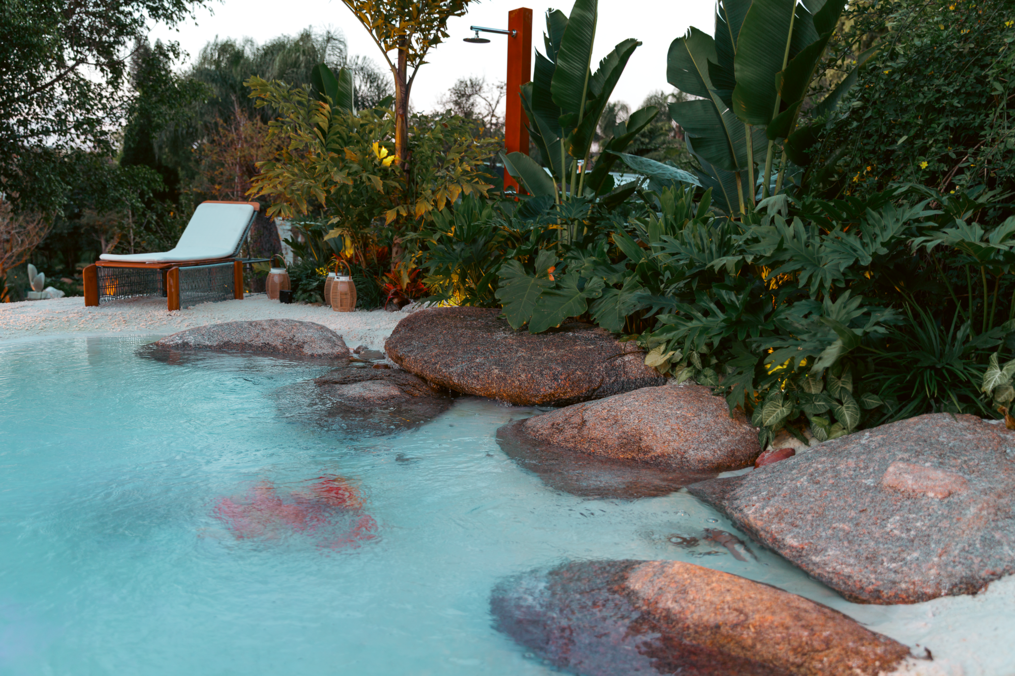 Projeto paisagístico tropical contemporâneo com piscina naturalística estilo praia, cujas bordas são compostas por grandes rochas graníticas avermelhadas e faixa de areia branca, criando uma atmosfera de lagoa natural. O entorno é densamente vegetado com espécies tropicais de folhagem exuberante como Strelitzia nicolai, Philodendron bipinnatifidum e Monstera deliciosa, conferindo privacidade e imersão na natureza. A área de estar conta com espreguiçadeira em madeira nobre e ducha externa em pila, estilo Tropical, Moderno, elementos Piscina de borda natural estilo praia com água cristalina azul-turquesa, Pedras graníticas naturais de grande porte formando borda orgânica da piscina, Areia branca ao redor da piscina simulando praia, Espreguiçadeira em madeira com estofado branco, Ducha externa em estrutura de madeira/aço corten com chuveirão, Vaso decorativo cerâmico estilo barril ao lado da espreguiçadeira, Maciço vegetal tropical denso ao redor da piscina, plantas Bananeira-ornamental, Costela-de-adão, Filodendro, Helicônia, Cróton, Guaimbê, Árvore-de-médio-porte não identificada com precisão, Palmeira-leque ou espécie arbustiva ao fundo, ambiente relaxante, sofisticado, acolhedor, localização piscina, jardim, quintal, área medio, estruturas iluminacao, cores azul-turquesa, verde-escuro, verde-claro, marrom-avermelhado, branco, bege, terracota, cinza-granito, piso pedra, brita, privacidade cerca_viva, água piscina