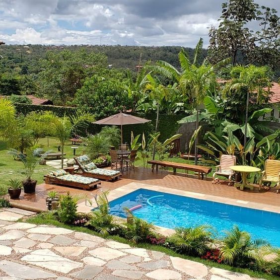 Projeto paisagístico de estilo tropical em propriedade ampla com piscina retangular emoldurada por generoso deck de madeira cumaru, circundada por maciços volumosos de bananeiras-ornamentais, heliconias e palmeiras-areca que criam um envolvente cenário exuberante com múltiplas camadas de verde. O jardim se desenvolve em patamares, com piso de pedra São Tomé irregular fazendo a transição entre o gramado esmeralda elevado e a área de lazer da piscina, enquanto canteiros de forração com bromélias, , estilo Tropical, Classico, elementos Piscina retangular revestida em azulejo azul, Deck de madeira cumaru ao redor da piscina, Espreguiçadeiras de madeira com almofadas estampadas tropicais, Ombrelone de centro em tom marrom, Mesa alta com banquetas em madeira sob o ombrelone, Banco comprido de madeira tipo rústico, Cadeira dobrável de madeira amarela, Cadeira dobrável de madeira verde, Mesa lateral redonda amarela, Vasos cerâmicos em tons escuros com plantas, Piso de pedra São Tomé irregular tipo pisante, Bóia infantil flutuando na piscina, Gramado amplo em nível elevado, Canteiros com bordaduras de plantas baixas, plantas Bananeira, Bananeira-ornamental, Palmeira-areca, Helicônia, Moreia, Grama-esmeralda, Agapanto, Cróton, Dracena-vermelha, Lírio-do-brejo, Samambaia, Clúsia, Ixora, Singônio, Alpínia, Filodendro, Palmeira-leque, Bromélia, ambiente relaxante, acolhedor, localização piscina, quintal, jardim, área grande, estruturas banco, cores verde-escuro, verde-claro, azul-piscina, marrom-madeira, amarelo, bege-pedra, branco, rosa, piso grama, deck, pisantes, pedra, privacidade cerca_viva, água piscina