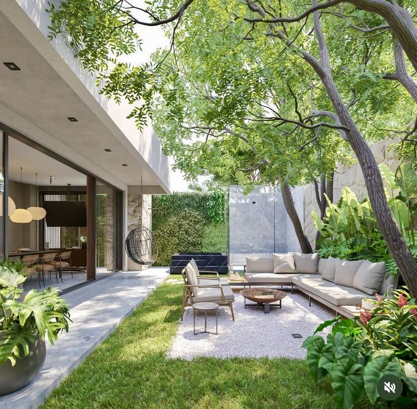 Jardim interno de residência contemporânea com forte integração indoor-outdoor, onde um generoso vão de esquadrias de vidro conecta a sala de jantar a um pátio ajardinado com lounge ao ar livre sob a copa de árvores de médio porte. O paisagismo explora volumetria tropical exuberante com folhagens largas de costela-de-adão, heliconias e bananeiras-ornamentais contrastando com a sobriedade do concreto aparente, muros de pedra natural e um jardim vertical que suaviza os limites do terreno. A área d, estilo Moderno, Tropical, elementos Sofá em L modular com estofado bege claro, Poltrona de madeira com almofada bege, Mesa de centro redonda em madeira rústica, Cadeira suspensa tipo balanço em estrutura de metal preto (egg chair), Luminárias pendentes esféricas em tom creme/off-white, Mesa de jantar com cadeiras em madeira e rattan visíveis no interior, Porta de correr ampla em vidro com esquadria de madeira escura, Parede de pedra natural em tons de cinza e bege, Jardim vertical com trepadeiras no muro de fundo, Parede/painel em concreto aparente como elemento escultórico, Vaso grande em concreto cinza com filodendro, Piso em área de brita branca sob o lounge, Forro/laje em concreto aparente branco com spots embutidos, Banco ou base em pedra preta (mármore negro) junto ao muro verde, plantas Guapuruvu, Helicônia, Bananeira-ornamental, Costela-de-adão, Filodendro, Jiboia, Pacová, Hera, Singônio, Grama-esmeralda, Taioba-ornamental, Asplenio, Unha-de-gato, ambiente relaxante, moderno, sofisticado, acolhedor, localização jardim, quintal, área medio, estruturas iluminacao, banco, cores branco, bege, cinza, verde, madeira natural, preto, off-white, creme, piso grama, brita, porcelanato, privacidade muro_verde, cerca_viva, água nenhum