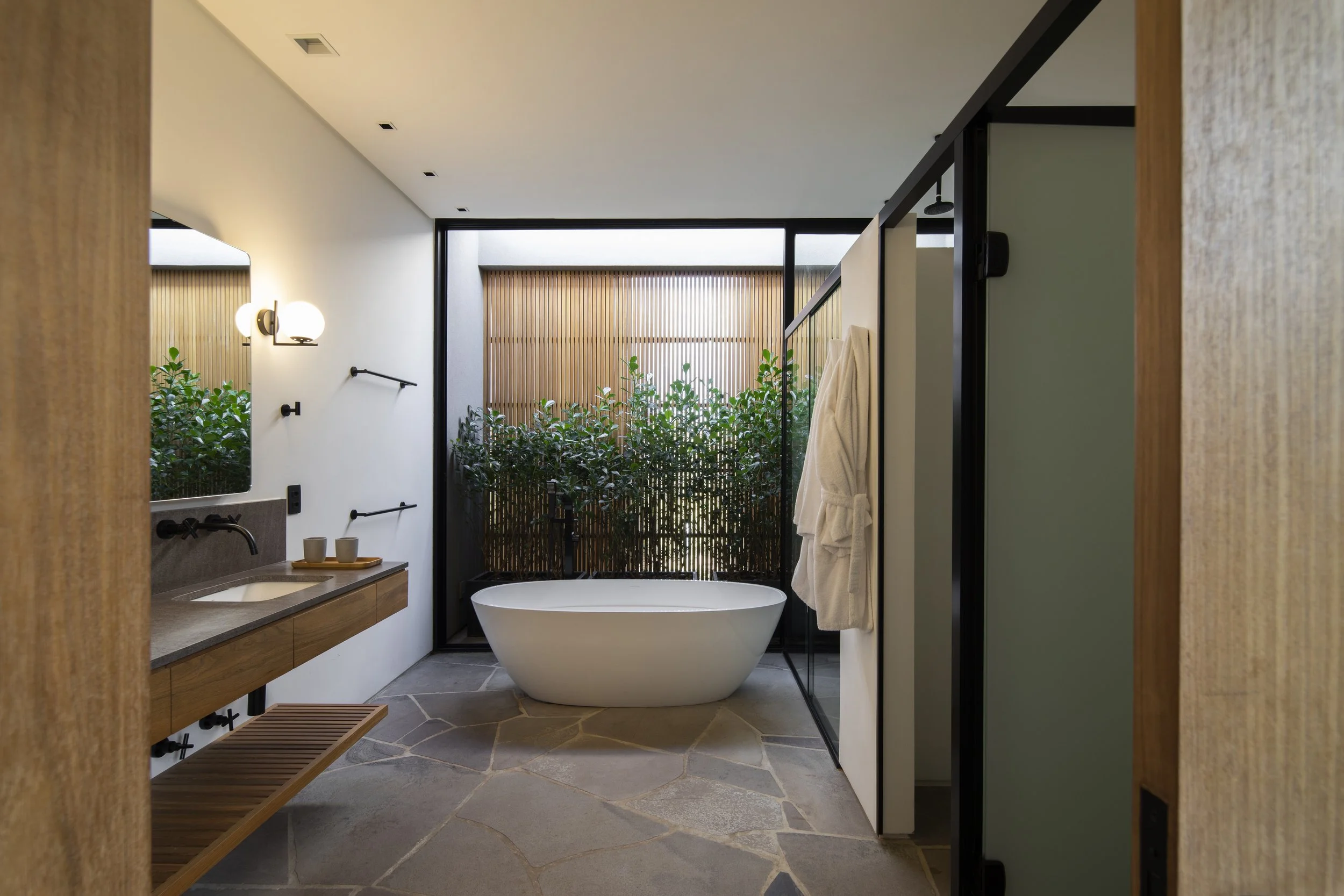 Banheiro contemporâneo com forte influência biofílica, onde uma generosa esquadria piso-teto em alumínio preto enquadra um jardim de inverno composto por maciço denso de clusias e painel ripado em madeira natural, criando uma cortina vegetal que garante privacidade e conexão com a natureza. O piso em pedra São Tomé irregular de formato orgânico (crazy paving) em tons de cinza contrasta com a bancada suspensa em madeira freijó e a banheira oval freestanding branca, reforçando a atmosfera de spa. , estilo Moderno, Tropical, elementos banheira oval freestanding branca, bancada em pedra natural com gabinete em madeira freijó, piso em pedra São Tomé irregular (crazy paving), painel ripado vertical em madeira natural (freijó ou cumaru), esquadria em alumínio preto piso-teto com vidro fixo, arandela globo esférico com acabamento dourado, metais de parede em preto fosco (torneira, porta-toalha, ducha), deck ripado em madeira sob a bancada, espelho horizontal embutido sobre a bancada, roupão branco pendurado em gancho, box com divisória em vidro temperado com perfil preto, spot embutido no forro de gesso, plantas Clusia, Bambu-mossô, ambiente relaxante, moderno, sofisticado, acolhedor, localização corredor, jardim, área pequeno, estruturas iluminacao, cores branco, cinza, preto fosco, madeira natural (tons de carvalho/freijó), verde folhagem, piso pedra, privacidade cerca_viva, água nenhum