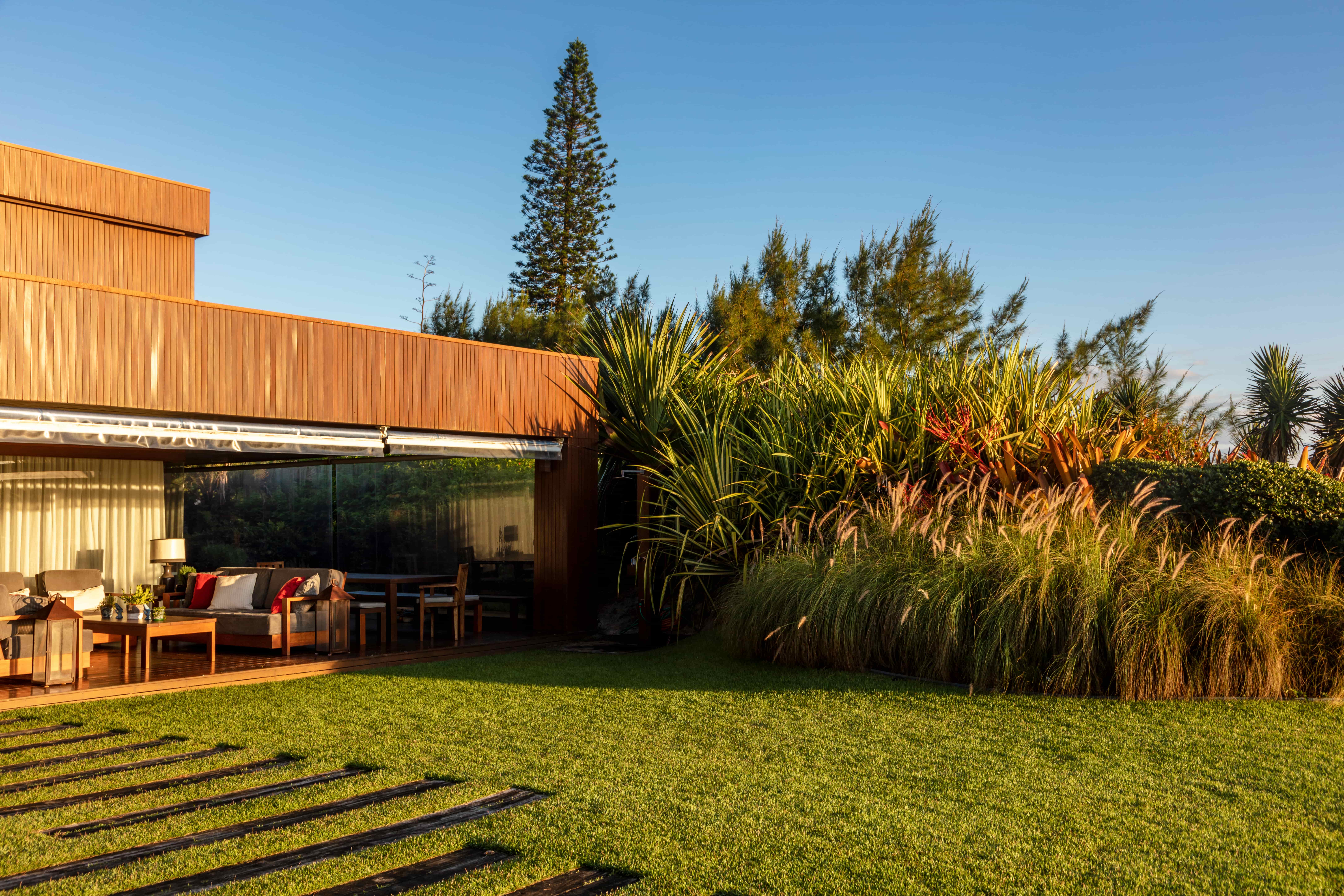 Projeto paisagístico contemporâneo de residência litorânea com volumetria em madeira cumaru, onde o generoso gramado de esmeralda conecta o deck da varanda a um exuberante maciço tropical composto por capins ornamentais, formios, dracenas e pandanos que criam uma cortina vegetal de alta densidade e textura variada. Os pisantes de madeira embutidos na grama conduzem o percurso de forma orgânica até o living externo protegido por vidro temperado e toldo retrátil, com mobiliário elegante em tons ne, estilo Moderno, Tropical, elementos Deck em madeira de lei com acabamento natural, Pisantes de madeira embutidos na grama, Painel de fechamento em vidro temperado com cortina branca, Fachada revestida em réguas de madeira cumaru vertical, Mobiliário externo em madeira com estofados cinza e almofadas vermelhas e brancas, Mesa lateral em madeira maciça, Toldo retrátil metálico sobre a varanda, Maciço vegetal denso como barreira visual natural, plantas Capim-do-texas, Dracena-vermelha, Pandano, Pinheiro-de-Norfolk, Casuarina, Helicônia, Formio, Grama-esmeralda, Iuca, Bromélia-imperial, ambiente relaxante, moderno, sofisticado, acolhedor, localização jardim, varanda, quintal, área grande, estruturas iluminacao, cores Verde-esmeralda da grama, Dourado-mel da madeira, Vermelho-bordô dos capins e cordylines, Branco das cortinas e almofadas, Cinza dos estofados, Verde-escuro da vegetação tropical, Azul-celeste do céu, piso grama, deck, pisantes, privacidade cerca_viva, água nenhum