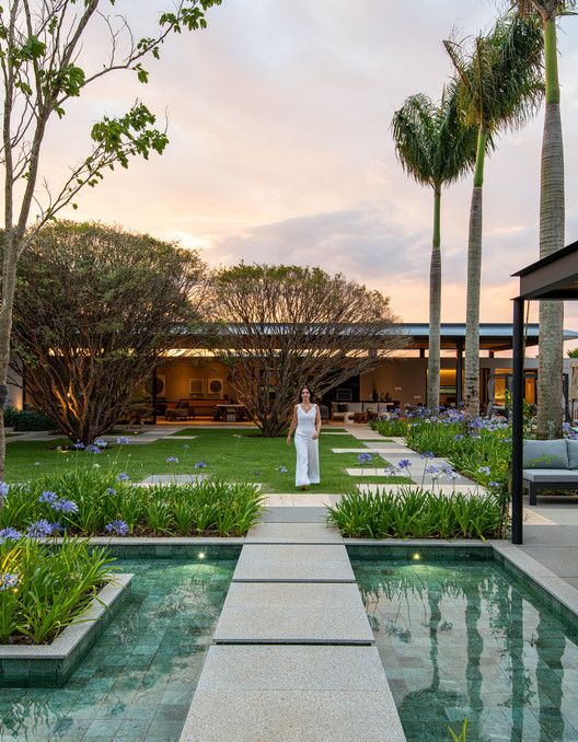 Projeto paisagístico de grande porte com linguagem contemporânea tropical, articulado em torno de um eixo central de pisantes de granito claro que atravessam um espelho d'água geométrico iluminado, conectando a área da piscina ao living aberto da residência ao fundo. Massas de agapantos em floração lilás emolduram os canteiros lineares junto aos espelhos d'água, enquanto exemplares adultos de jasmim-manga com troncos escultóricos e palmeiras-reais imponentes conferem verticalidade e caráter trop, estilo Moderno, Tropical, elementos Espelho d'água retangular com pisantes de granilha/granito claro, Pisantes retangulares de pedra clara sobre a grama, Iluminação subaquática embutida no espelho d'água, Pergolado metálico escuro com estrutura contemporânea à direita, Cobertura horizontal com telha metálica ao fundo integrando área de estar, Mobiliário lounge externo sob pergolado, Canteiros lineares com bordadura junto ao espelho d'água, Piso de pedra natural nos passeios, plantas Agapanto, Palmeira-real, Plumeria / Jasmim-manga, Grama-esmeralda, Moreia, Lírio-do-Nilo, ambiente sofisticado, moderno, relaxante, localização jardim, piscina, entrada, área grande, estruturas pergolado, iluminacao, cores verde, lilás, branco, cinza claro, marrom, preto, piso grama, pisantes, pedra, privacidade cerca_viva, água espelho_dagua, piscina