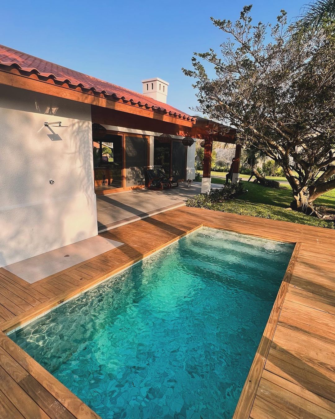 Projeto paisagístico de residência em estilo contemporâneo com influência rústica, destacando-se uma elegante piscina retangular com água cristalina turquesa emoldurada por generoso deck em madeira nobre, que se integra harmoniosamente à varanda coberta por pergolado de madeira com telhado colonial em barro. O jardim conta com um exemplar imponente de figueira de tronco escultórico posicionada estrategicamente ao lado da piscina, criando sombra natural e um ponto focal orgânico, complementada po, estilo Moderno, Tropical, elementos Piscina retangular com borda em deck de madeira, Deck de madeira natural tipo cumaru ou teca ao redor da piscina, Ducha externa em aço inox fixada na parede lateral da casa, Pergolado em madeira de lei com cobertura parcial em telha colonial, Telhado colonial com telhas de barro em tom terracota, Chaminé em alvenaria branca, Portas e janelas amplas em vidro com esquadrias de madeira, Parede externa em alvenaria com acabamento branco, Piso da varanda em porcelanato ou cimentício, Gramado amplo e bem aparado ao redor do jardim, plantas Figueira-de-jardim, Grama-esmeralda, Palmeira, Arbusto ornamental não identificado, ambiente relaxante, sofisticado, acolhedor, localização piscina, jardim, varanda, quintal, área medio, estruturas pergolado, iluminacao, cores Turquesa da água da piscina, Marrom-dourado do deck de madeira, Branco das paredes externas, Terracota das telhas coloniais, Verde intenso do gramado e folhagens, Azul claro do céu, piso deck, grama, porcelanato, privacidade nenhum, água piscina