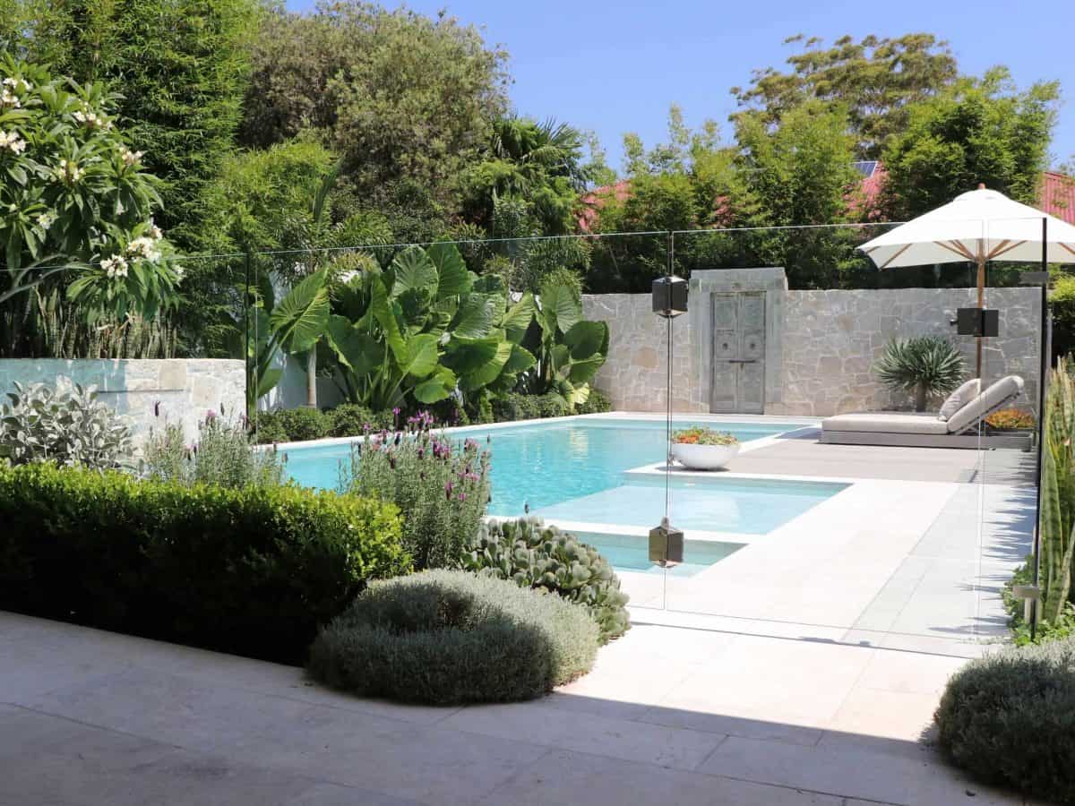 Projeto paisagístico contemporâneo integrado a uma piscina retangular com spa embutido, delimitado por elegante cerca de vidro temperado sem moldura que permite total visibilidade do jardim tropical exuberante ao fundo. A composição vegetal combina espécies tropicais de grande porte como taro-gigante e bambu com arbustos esculturais de buxinho topiado e massas de lavanda e santolina em tons prateados, criando camadas de textura e volume contra o muro de pedra natural rústica. O deck em porcelana, estilo Moderno, Tropical, elementos Piscina retangular com borda infinita em tom azul-claro, Spa/jacuzzi integrado à piscina em nível rebaixado, Deck em porcelanato bege claro ao redor da piscina, Muro de pedra natural em tons de cinza e bege, Cerca de vidro temperado frameless ao redor da piscina, Espreguiçadeira estofada em tom neutro com ombrelone branco, Porta antiga decorativa de madeira rústica no muro de pedra, Vasos decorativos brancos no deck, Caixas de som embutidas no muro de pedra, plantas Alamanda, Taro-gigante, Bambu, Buxinho, Iúca, Lavanda, Rosa-do-deserto ou Plumeria, Echevéria, Cacto-candelabro, Santolina, Alecrim, Agave, Murta, Verbena, ambiente relaxante, sofisticado, moderno, localização piscina, jardim, quintal, área grande, estruturas iluminacao, cores verde-escuro, verde-claro, branco, bege, azul-claro, cinza, lilás, rosa, piso porcelanato, pedra, privacidade cerca_viva, muro_verde, água piscina