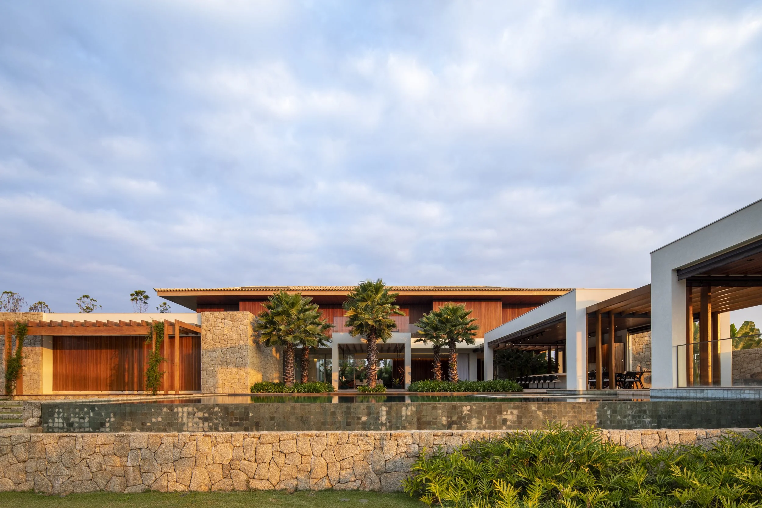 Projeto paisagístico de grande escala integrado a uma residência contemporânea com influências tropicais, onde palmeiras-leque esculturais são posicionadas como protagonistas em canteiros elevados sobre muros de pedra natural, criando uma composição vertical que dialoga com as linhas horizontais da arquitetura. O espelho d'água linear revestido em pedra escura percorre toda a extensão frontal da casa, refletindo o volume arquitetônico e conferindo sofisticação ao conjunto, enquanto o extenso gra, estilo Moderno, Tropical, elementos Muro de pedra natural irregular em tons terrosos, Espelho d'água linear com revestimento em pedra escura, Pergolado de madeira sobre estrutura lateral esquerda, Fachada com painéis de madeira cumaru e paredes brancas, Telhado colonial com telhas cerâmicas, Pilares brancos de concreto aparente, Canteiros elevados com bordadura em pedra, Gramado plano extenso no primeiro plano, Trepadeira em crescimento sobre o pergolado lateral, plantas Palmeira-leque-da-china, Palmeira-bismarckia, Bromélia-imperial, Agave, Moréia, Grama-esmeralda, Unha-de-gato, Palmeira-buriti, Pândano, Clúsia, Helicônia, ambiente sofisticado, moderno, relaxante, localização fachada, jardim, piscina, entrada, área grande, estruturas pergolado, iluminacao, cores Verde-escuro, Verde-claro, Bege-areia, Marrom-madeira, Branco, Cinza-pedra, Terracota, piso grama, pedra, privacidade muro_verde, cerca_viva, água espelho_dagua