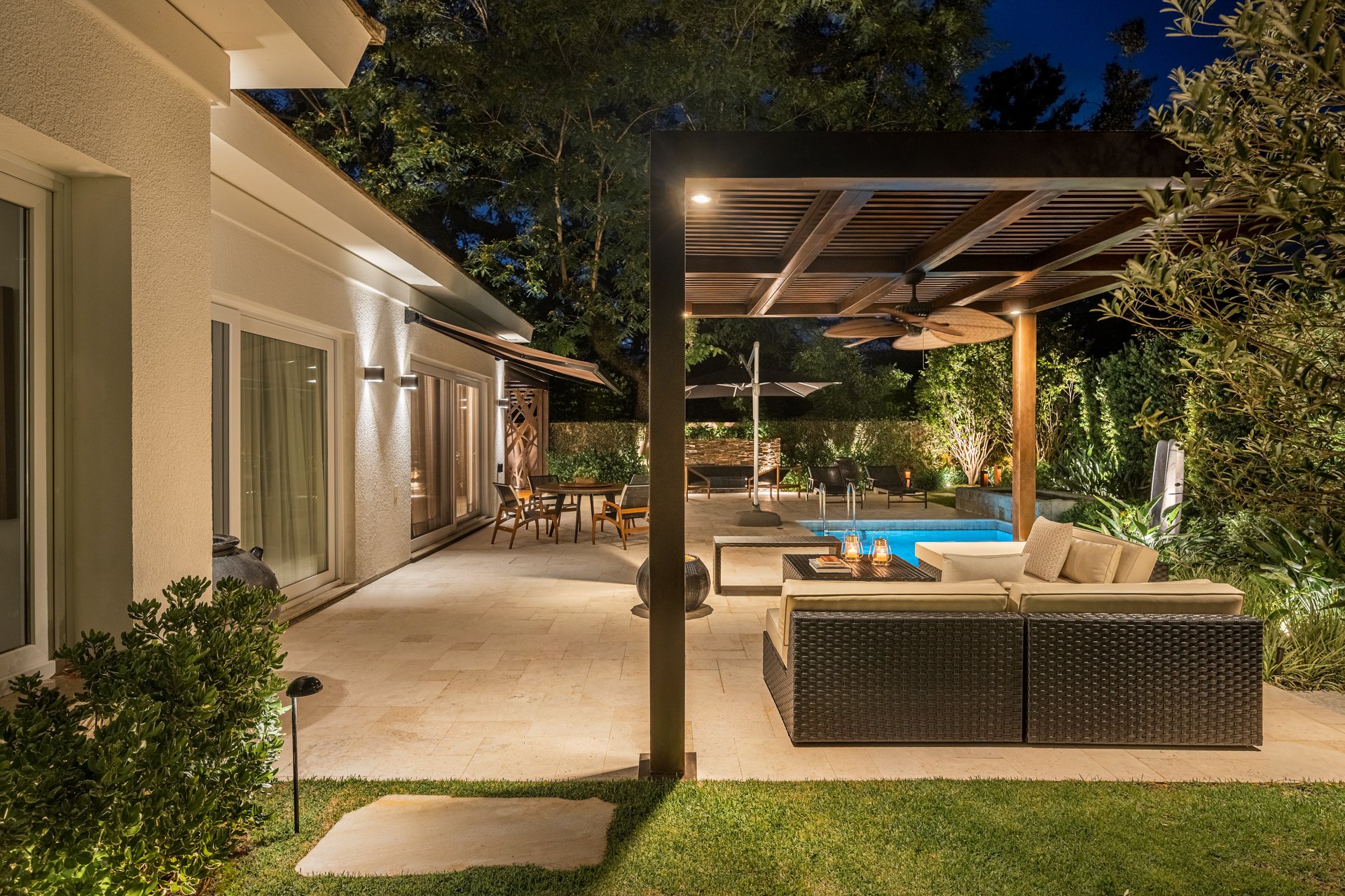 Projeto paisagístico de alto padrão em área externa residencial que integra elegantemente um lounge coberto por pergolado metálico com forro em madeira, área de jantar ao ar livre e piscina iluminada, tudo conectado por amplo piso em travertino romano. A vegetação tropical e mediterrânea — com oliveiras, buxinhos topiados, trepadeiras e massivos arbustivos — compõe uma cerca viva natural que garante privacidade total, enquanto a iluminação cênica com balizadores, spots de piso e wall washers cri, estilo Moderno, Tropical, elementos Sofá modular em fibra sintética com almofadas em tom cru, Mesa de centro com lanternas decorativas de vidro e metal, Pergolado em estrutura metálica preta com cobertura em réguas de madeira, Ventilador de teto com pás em madeira estilo colonial, Piscina com borda infinita e iluminação subaquática azul, Mesa de jantar retangular em madeira com cadeiras, Ombrelone preto articulável, Espreguiçadeiras ao redor da piscina, Toldo retrátil fixado na fachada da casa, Iluminação embutida em balizadores no jardim, Iluminação de destaque tipo wall washer na fachada, Pisantes retangulares em pedra natural sobre a grama, Piso em travertino romano na área externa, Treliça decorativa em madeira na parede da casa, Spot de piso para iluminação de árvores e arbustos, plantas Buxinho, Oliveira, Murta, Palmeira-leque, Jasmim-estrela, Unha-de-gato, Agave, Grama-esmeralda, Pleomele, Podocarpo, Helicônia, Clúsia, ambiente relaxante, moderno, sofisticado, acolhedor, localização jardim, piscina, quintal, varanda, área grande, estruturas pergolado, iluminacao, cores bege, marrom-escuro, verde, branco, preto, azul-piscina, cru, amadeirado, piso grama, pisantes, pedra, privacidade cerca_viva, muro_verde, água piscina