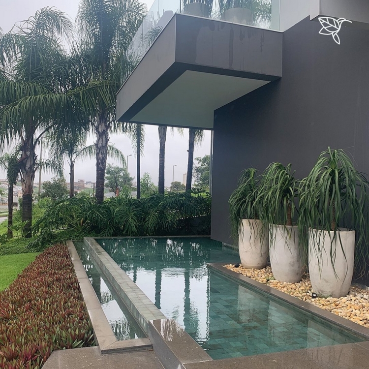 Projeto paisagístico contemporâneo integrado à arquitetura minimalista, onde uma piscina retangular de pastilhas esmeraldas cria um espelho d'água que reflete a volumetria do balanço em concreto e vidro da varanda superior. O paisagismo tropical é composto por palmeiras-reais imponentes ao fundo, patas-de-elefante em vasos de concreto escultural sobre seixos brancos, e um maciço denso de bromélias neoregélias avermelhadas e samambaias que suavizam a transição entre o mineral e o vegetal. A palet, estilo Moderno, Tropical, elementos Piscina retangular com borda em pedra São Tomé, Vasos de concreto grandes em formato cônico, Seixos rolados brancos decorativos ao redor dos vasos, Canteiro elevado lateral com bromélias e forrações, Varanda em balanço com guarda-corpo de vidro temperado, Parede revestida em granito ou porcelanato cinza escuro, Piso de pedra natural ao redor da piscina, plantas Palmeira-real, Pata-de-elefante, Palmeira-areca, Bromélia-imperial, Neoregélia, Samambaia-de-metro, Helicônia, ambiente sofisticado, moderno, relaxante, localização piscina, fachada, jardim, área medio, estruturas iluminacao, cores verde-escuro, verde-claro, cinza-chumbo, branco, bege, vermelho-vinho, esmeralda, piso pedra, brita, privacidade cerca_viva, água piscina, espelho_dagua