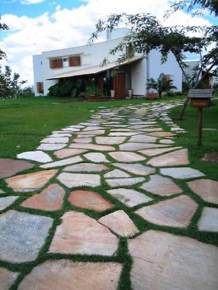 Projeto paisagístico de residência contemporânea com amplo jardim frontal, destacando-se um generoso caminho de pedras naturais irregulares tipo São Tomé em tons rosados e acinzentados, assentadas com juntas vivas de grama-esmeralda que criam um efeito orgânico e acolhedor. A vegetação tropical composta por bananeiras-ornamentais, palmeiras e árvores de médio porte como a sibipiruna emoldura a fachada branca e minimalista, cujas esquadrias em madeira nobre conferem calor e sofisticação ao conjun, estilo Moderno, Tropical, elementos Caminho de pedras naturais irregulares tipo São Tomé com juntas de grama, Placa de sinalização em madeira de lei sobre poste rústico, Cobertura/toldo em lona ou tecido tensionado sobre a varanda, Esquadrias em madeira maciça com venezianas em tom mogno, Porta principal em madeira de lei maciça com vidro, Fachada em alvenaria com acabamento branco liso, Gramado amplo e bem aparado em ambos os lados do caminho, plantas Grama-esmeralda, Bananeira-ornamental, Palmeira-real, Sibipiruna, Grama-amendoim, ambiente acolhedor, moderno, sofisticado, localização entrada, fachada, jardim, área grande, estruturas iluminacao, nenhum, cores Verde-esmeralda do gramado, Rosa-arenoso e cinza das pedras São Tomé, Branco da fachada, Marrom-mogno das esquadrias e portas, Bege do toldo, Verde-escuro das folhagens tropicais, piso pedra, grama, pisantes, privacidade nenhum, água nenhum