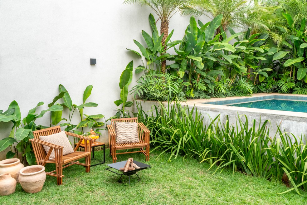 Projeto paisagístico de quintal compacto com forte identidade tropical brasileira, onde um maciço exuberante de bananeiras-ornamentais, heliconias, filodendros e palmeiras-fênix cria uma cortina verde densa que garante privacidade e ambiência de refúgio urbano junto a uma piscina elevada em concreto aparente. A área de estar é composta por mobiliário artesanal em bambu com almofadas em fibras naturais, acompanhado de uma fogueira portátil em metal e vasos de cerâmica artesanal, tudo disposto sob, estilo Tropical, Moderno, elementos Conjunto de estar em bambu com dois bancos e mesa lateral, Almofadas decorativas em tons neutros e trançado natural, Fogueira portátil de mesa em metal preto (fire pit), Vasos de cerâmica rústica em tamanhos variados, Piscina elevada com borda em concreto aparente e coping em pedra natural, Muro rebocado e pintado de branco, Arandela preta na parede para iluminação externa, Bandeja com copos de suco na mesa lateral, Canteiro elevado de concreto junto ao muro, plantas Bananeira-ornamental, Helicônia, Palmeira-fênix, Moréia, Filodendro, Lírio-do-brejo, Palmeira-areca, Costela-de-adão, Capim-do-texas, Samambaia, Grama-esmeralda, ambiente relaxante, acolhedor, sofisticado, localização quintal, piscina, jardim, área pequeno, estruturas banco, iluminacao, cores verde-escuro, verde-claro, branco, bege, terracota, preto, azul-piscina, cinza-concreto, piso grama, privacidade muro_verde, cerca_viva, água piscina