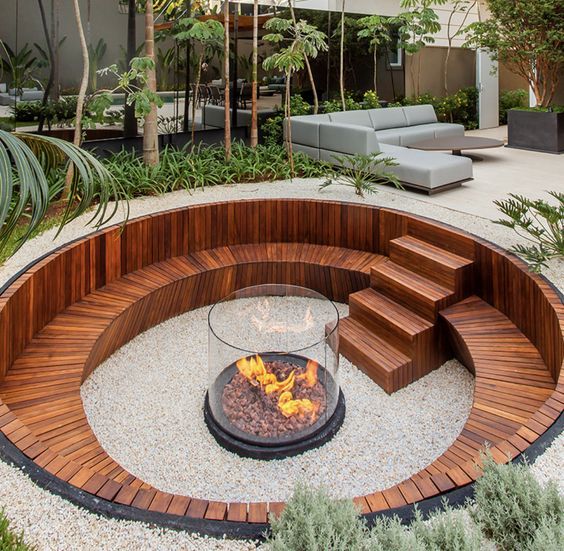 Sunken garden contemporâneo com banco circular em madeira de lei ripada, rebaixado em relação ao nível do jardim principal, centrado em uma elegante fogueira de vidro temperado sobre base metálica preta com cascalho vulcânico. O piso em brita branca compactada contrasta com o tom quente da madeira, enquanto canteiros perimetrais com vegetação tropical — dracenas, palmeiras-leque e forrações como liriopes — criam um envoltório verde exuberante que garante privacidade e aconchego. Ao fundo, a área, estilo Moderno, Tropical, elementos Fogueira central de vidro temperado com base metálica preta sobre cascalho vulcânico, Banco curvo embutido em madeira de lei (ipê ou cumaru) com acabamento ripado, Escada de acesso em madeira ripada com 5 degraus integrados ao banco circular, Borda metálica escura delimitando o sunken garden, Piso de brita branca compactada preenchendo a área rebaixada, Sofá modular cinza claro em área de estar elevada ao fundo, Canteiros perimetrais elevados com vegetação tropical, Mesa lateral em vidro transparente junto ao banco curvo, plantas Dracena-de-madagascar, Palmeira-leque, Moreia, Liriope, Barba-de-serpente, Alecrim, Flamboyanzinho, Helicônia, Singônio, Aspargo-pluma, ambiente relaxante, moderno, acolhedor, sofisticado, localização jardim, quintal, área medio, estruturas banco, iluminacao, cores marrom-escuro, branco, verde-escuro, verde-claro, cinza-claro, preto, laranja-fogo, piso brita, deck, privacidade cerca_viva, água nenhum