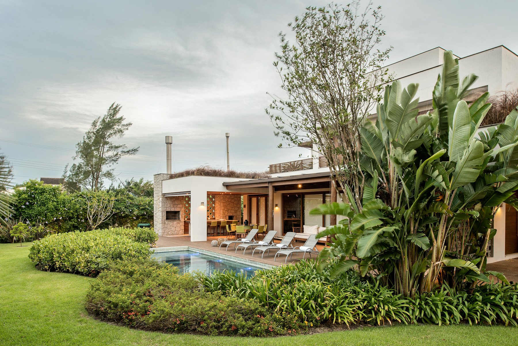 Projeto paisagístico tropical contemporâneo integrado a uma residência de linhas retas e volumetria escalonada, onde massas arbustivas orgânicas de ixoras, moréias e liriopes contornam um generoso gramado de esmeralda que emoldura a piscina retangular posicionada junto à área gourmet com churrasqueira em pedra. Estrelítzias-gigantes e bananeiras-ornamentais conferem verticalidade e exuberância tropical ao canto direito, enquanto frangipanes e casuarinas ao fundo criam um segundo plano volumétric, estilo Moderno, Tropical, elementos Piscina retangular com borda em pedra natural, Espreguiçadeiras em alumínio com tela sling cinza (4 unidades), Churrasqueira revestida em pedra rústica com chaminé metálica, Cobertura em pergolado com estrutura metálica e viga de madeira, Telhado verde/jardim sobre laje na cobertura da edícula, Parede decorativa com elementos vazados em cerâmica, Iluminação embutida em arandelas nas paredes externas, Mesa de jantar com cadeiras amarelas na área gourmet, Mureta branca com acabamento em massa acrílica, Piso em deck de pedra ao redor da piscina, Gramado extenso com forração em massas arbustivas orgânicas, plantas Helicônia, Bananeira-ornamental, Estrelítzia-gigante, Moreia, Ixora, Pingo-de-ouro, Frangipani, Casuarina, Palmeira-areca, Grama-esmeralda, Agapanto, Clúsia, Líriope, Trapoeraba-roxa, ambiente relaxante, moderno, sofisticado, localização jardim, piscina, varanda, quintal, área grande, estruturas pergolado, churrasqueira, iluminacao, cores verde-escuro, verde-claro, branco, cinza, bege, amarelo, terracota, marrom, piso grama, pedra, privacidade cerca_viva, água piscina