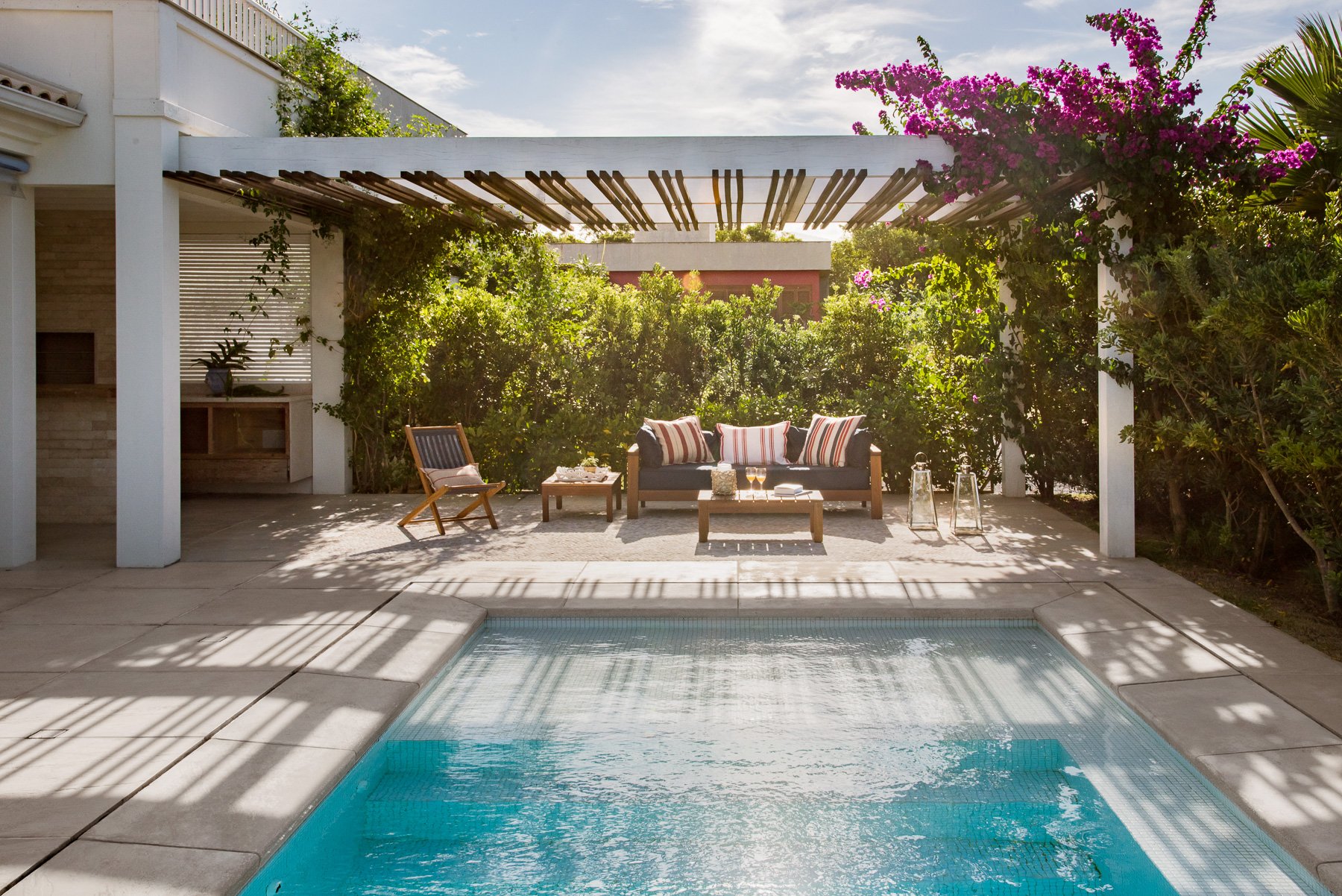 Projeto paisagístico de estilo moderno-tropical que integra uma piscina retangular com revestimento em pastilha azul-turquesa a uma área de estar coberta por elegante pergolado de concreto branco com travessas de madeira, cercada por densa vegetação tropical que garante total privacidade. A exuberante primavera (Bougainvillea spectabilis) em plena floração magenta no canto superior direito contrasta com o verde intenso da cerca viva de murtas e arbustos ao fundo, enquanto uma palmeira-leque adic, estilo Moderno, Tropical, elementos piscina retangular com revestimento em pastilha azul-turquesa, pergolado de concreto branco com travessas de madeira, sofá de área externa com almofadas listradas em tons de terracota e branco, cadeira dobrável de madeira teca, mesa de centro baixa de madeira, mesa lateral de madeira, par de lanternas decorativas de vidro e metal, piso de placas de concreto ao redor da piscina, tapete de área externa sob o mobiliário, taças de vinho sobre a mesa de centro, cerca viva densa formada por arbustos ao fundo, pilares brancos de alvenaria sustentando o pergolado, churrasqueira ou bancada de apoio em pedra natural à esquerda, plantas Primavera, Palmeira-leque, Murta, Jasmim-manga, Pitosporo, Ficus, Hibisco, ambiente relaxante, sofisticado, acolhedor, localização piscina, quintal, jardim, área medio, estruturas pergolado, churrasqueira, iluminacao, cores branco, azul-turquesa, verde, magenta, terracota, bege, madeira natural, piso porcelanato, brita, privacidade cerca_viva, água piscina