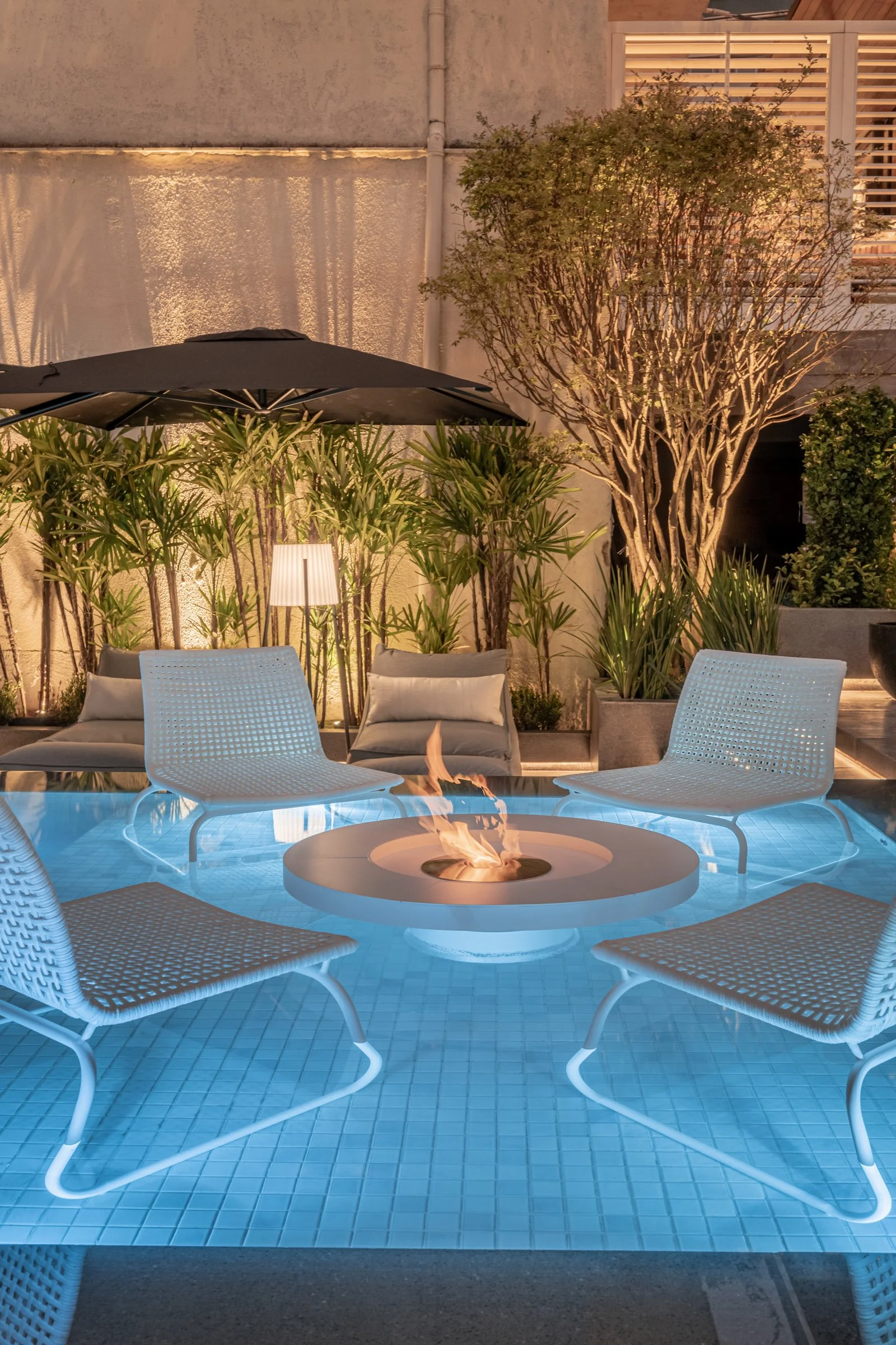 Projeto paisagístico contemporâneo integrado a uma área de piscina urbana, onde um elegante conjunto de mobiliário em fibra sintética branca com mesa de lareira ecológica central é posicionado dentro da lâmina d'água rasa, criando um lounge aquático sofisticado. O paisagismo tropical compõe o entorno com maciços de ráfis (Rhapis excelsa) retroiluminados por spots de uplighting, uma jasmim-manga (Plumeria rubra) de troncos escultóricos como ponto focal e topiaria de buxinho, todos emoldurados por, estilo Moderno, Tropical, elementos Piscina com revestimento em pastilha azul clara, Conjunto de cadeiras em fibra sintética branca dentro da piscina (deck molhado/pool lounge), Mesa central com lareira ecológica a etanol embutida, Ombrelone lateral preto de grande porte, Espreguiçadeira/daybed com almofadas em tom cinza, Luminária de piso com cúpula branca estilo contemporâneo, Vasos em concreto/cimento com acabamento cinza, Muro de concreto aparente ao fundo, Janela com veneziana horizontal branca no pavimento superior, Iluminação cênica direcionada nas plantas (uplighting), plantas Ráfis, Dracena-de-madagascar, Jabuticabeira ou árvore de tronco escultural — possivelmente Jasmim-manga, Buxinho, ambiente sofisticado, moderno, relaxante, localização piscina, quintal, área medio, estruturas iluminacao, cores azul claro, branco, cinza, verde, preto, bege, laranja (fogo), piso porcelanato, privacidade cerca_viva, muro_verde, água piscina