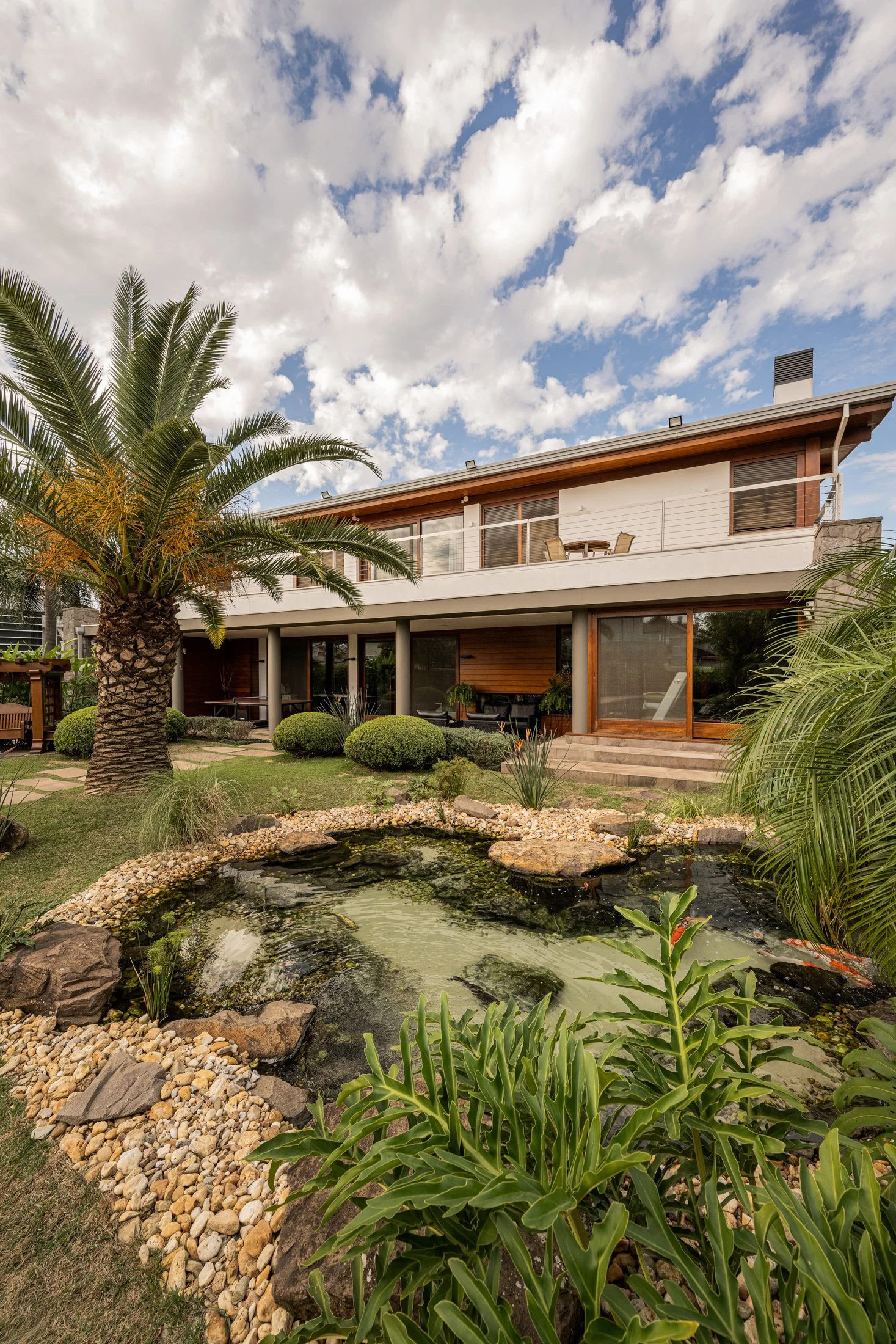 Projeto paisagístico de estilo tropical-contemporâneo integrado a uma residência moderna de dois pavimentos com amplo uso de madeira e vidro, destacando-se um lago ornamental naturalístico com bordas de pedras brutas de basalto e seixos rolados, que abriga plantas aquáticas e peixes ornamentais, criando um microecossistema no jardim frontal. A composição vegetal mescla uma imponente palmeira-fênix como ponto focal, maciços de buxinho topiados em formas esféricas que conferem ritmo e estrutura ao, estilo Moderno, Tropical, elementos Lago ornamental naturalístico com pedras naturais irregulares e seixos rolados, Bordadura de pedras brutas e seixos de rio em tons bege e ocre, Maciços arbustivos topiados em formato arredondado, Forração verde contínua em grama esmeralda bem mantida, Rochas rústicas de basalto dispostas de forma orgânica ao redor do lago, Casa contemporânea de dois pavimentos com varanda e acabamento em madeira de lei, Chaminé em alvenaria na cobertura, Pilaretes de concreto sustentando varanda superior, plantas Palmeira-fênix, Palmeira-areca, Buxinho, Guaimbê, Helicônia, Grama-esmeralda, Ninféia, Papiro-anão, Moreia, Clúsia, ambiente relaxante, sofisticado, moderno, acolhedor, localização jardim, fachada, quintal, área grande, estruturas iluminacao, churrasqueira, cores Verde-escuro, Verde-claro, Bege, Marrom-madeira, Branco, Ocre, Cinza-pedra, Vermelho pontual (helicônia/flores), piso grama, pedra, brita, pisantes, privacidade cerca_viva, água espelho_dagua