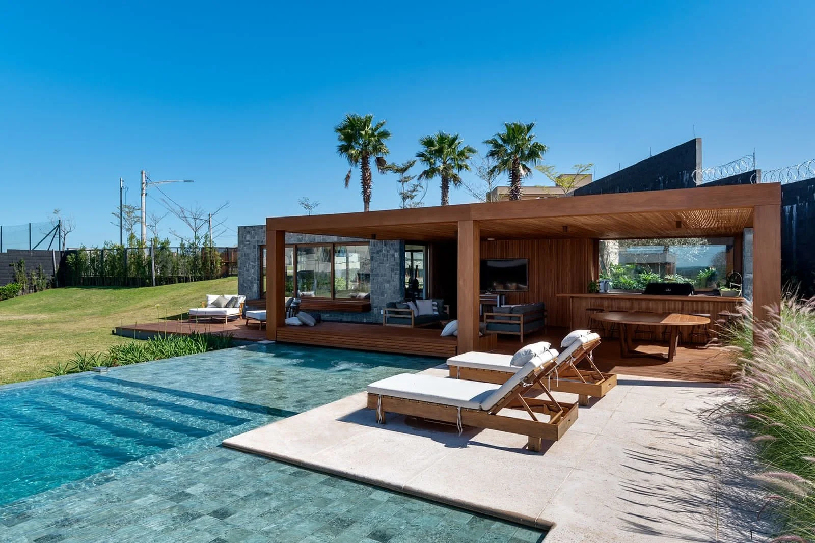 Projeto paisagístico contemporâneo de alto padrão integrado a uma residência horizontal com volumetria em madeira e pedra, onde a piscina de borda infinita com revestimento em pedra natural funciona como elemento central conectando o deck elevado, a área de espreguiçadeiras e o jardim gramado em desnível. A vegetação tropical composta por palmeiras-washingtônia em trio, capim-do-texas em maciços ornamentais e forração verde cria enquadramento visual sofisticado, enquanto o pergolado em madeira m, estilo Moderno, Tropical, elementos Piscina de borda infinita com revestimento em pedra natural verde, Deck em madeira cumaru elevado com área de estar, Espreguiçadeiras em madeira com estofado branco, Pergolado em madeira maciça com cobertura em bambu, Mesa redonda em madeira maciça com cadeiras para área gourmet, Sofás e poltronas externos com almofadas neutras, Piso em limestone/pedra natural bege ao redor da piscina, Muro perimetral escuro com cerca concertina no topo, Iluminação embutida na área coberta, TV externa na área de estar coberta, Postes de energia ao fundo indicando área suburbana, plantas Palmeira-washingtônia, Capim-do-texas, Grama-esmeralda, Moréia, Clúsia, Helicônia, Bambuzinho-de-jardim, ambiente relaxante, moderno, sofisticado, localização piscina, jardim, varanda, quintal, área grande, estruturas pergolado, churrasqueira, iluminacao, banco, cores marrom-madeira, bege-areia, verde-esmeralda, azul-turquesa, branco, cinza-pedra, preto, piso grama, deck, pedra, privacidade cerca_viva, muro_verde, água piscina