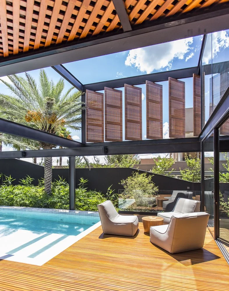Projeto paisagístico contemporâneo tropical que integra uma piscina retangular com borda infinita a um generoso deck de cumaru, coberto por um sofisticado pergolado de treliça de madeira ripada sustentado por estrutura metálica preta, complementado por painéis pivotantes tipo muxarabi que filtram a luz e garantem privacidade. A vegetação tropical exuberante com tamareiras imponentes, arbustos densos de clúsia e podocarpo cria um cinturão verde envolvente que contrasta com os muros pretos e confe, estilo Moderno, Tropical, elementos Pergolado com treliça de madeira ripada sobre estrutura metálica preta, Painéis pivotantes de madeira vazada tipo muxarabi, Deck de madeira cumaru em réguas longitudinais junto à piscina, Piscina retangular com borda infinita e revestimento azul claro, Duas poltronas de design contemporâneo em couro bege com almofadas brancas, Mesa lateral redonda em madeira maciça tipo toco, Fechamento em vidro temperado piso-teto com esquadria preta, Muro de alvenaria alto pintado de preto fosco como barreira de privacidade, Estrutura metálica em aço pintado preto com vigas aparentes, Guarda-corpo em vidro transparente junto ao deck da piscina, plantas Tamareira, Palmeira-fênix, Murta, Podocarpo, Helicônia, Clúsia, Viburno, Moréia, ambiente sofisticado, moderno, relaxante, localização piscina, jardim, quintal, área medio, estruturas pergolado, cores madeira natural, preto, bege, branco, verde, azul piscina, marrom, piso deck, privacidade cerca_viva, muro_verde, água piscina