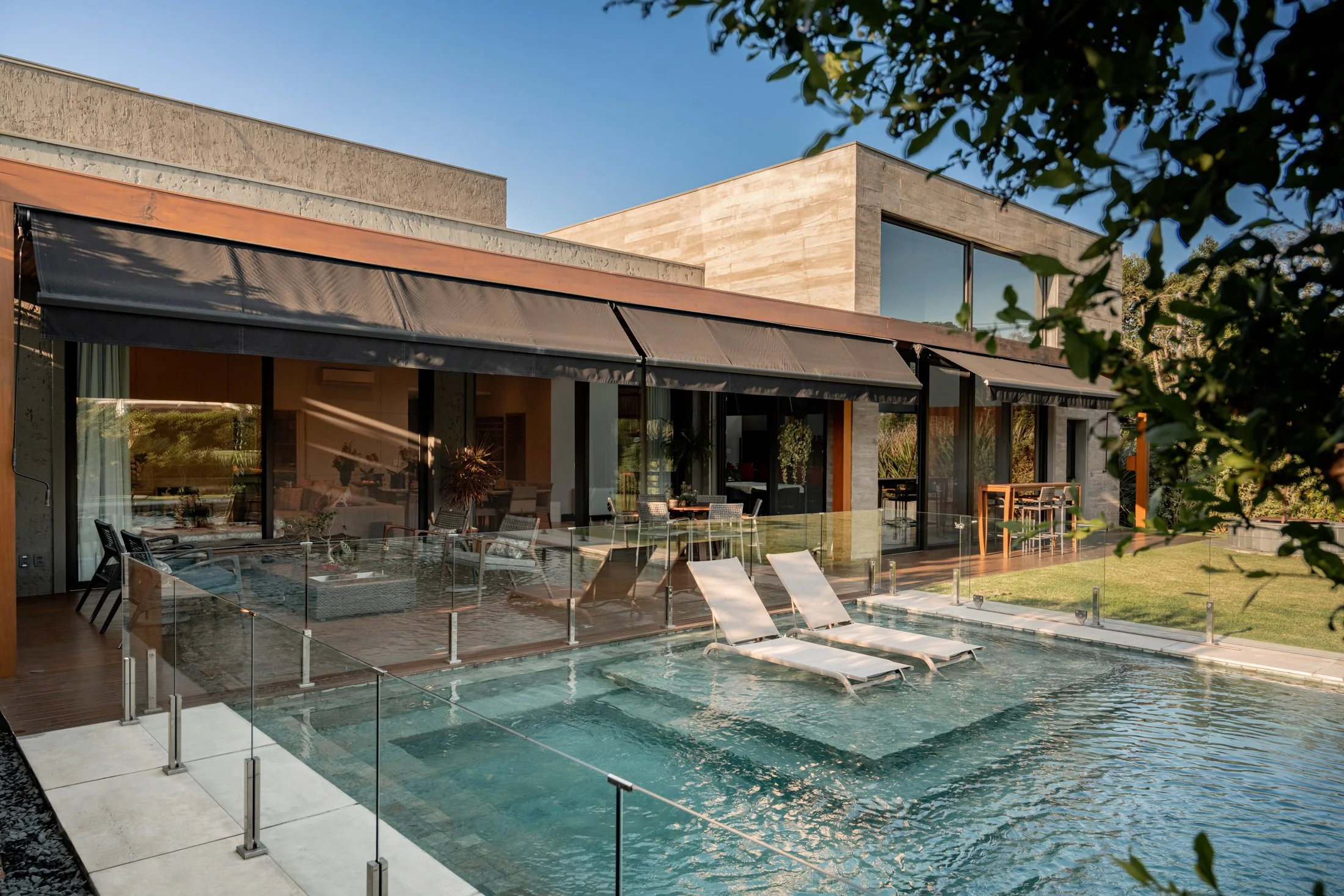Projeto paisagístico contemporâneo de alto padrão integrado a uma residência com volumetria marcante em travertino e aço corten, destacando-se a piscina com prainha (sun shelf) equipada com espreguiçadeiras submersas, emoldurada por guarda-corpo de vidro e deck de madeira nobre. A vegetação combina gramíneas ornamentais como capim-do-texas de folhagem arroxeada e capim-dos-pampas ao fundo, com massas arbustivas verdes que criam privacidade sem comprometer a linguagem minimalista. O conjunto harm, estilo Moderno, Tropical, elementos Piscina com borda infinita e prainha interna (sun shelf) com espreguiçadeiras dentro da água, Guarda-corpo de vidro temperado com colunas em aço inox escovado, Deck em madeira cumaru ao redor da piscina, Piso em porcelanato claro de grande formato na área de borda, Toldos retráteis em lona escura sobre a fachada envidraçada, Espreguiçadeiras de alumínio com tecido sling branco, Mesa de jantar externa em madeira com cadeiras, Esquadrias em vidro piso-teto com perfil preto, Fachada revestida em pedra travertino e aço corten, Gramado amplo lateral bem aparado, Iluminação embutida subaquática na piscina, plantas Capim-do-texas, Dracena-vermelha, Capim-dos-pampas, Grama-esmeralda, Mangueira ou árvore de médio porte nativa, Murta ou arbusto de cerca viva, Clúsia, ambiente sofisticado, moderno, relaxante, localização piscina, jardim, fachada, quintal, área grande, estruturas iluminacao, cores bege, cinza, verde, branco, marrom corten, azul água, preto, piso grama, deck, porcelanato, privacidade cerca_viva, água piscina