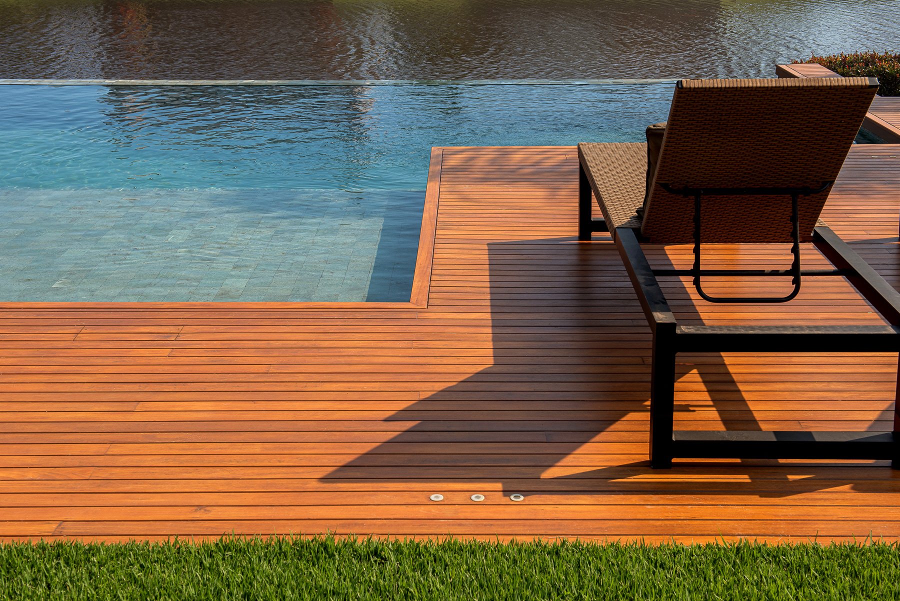 Projeto paisagístico contemporâneo de alto padrão centrado em uma piscina com borda infinita revestida em pedra natural de tonalidade azulada, ladeada por um amplo deck em madeira nobre (possivelmente cumaru ou ipê) com acabamento impecável e coloração alaranjada quente. O gramado de grama-esmeralda perfeitamente mantido faz a transição suave entre o deck e o restante do jardim, enquanto espreguiçadeiras em fibra sintética trançada e spots de LED embutidos no piso conferem funcionalidade e sofis, estilo Moderno, Tropical, elementos Deck em madeira cumaru com acabamento envernizado, Piscina com borda infinita e revestimento em pedra natural tom azulado, Espreguiçadeiras em fibra sintética marrom com estrutura metálica preta, Iluminação embutida no deck (spots de piso em LED), Gramado bem aparado junto ao deck, plantas Grama-esmeralda, ambiente relaxante, moderno, sofisticado, localização piscina, jardim, área grande, estruturas iluminacao, cores Marrom avermelhado do deck, Azul turquesa da piscina, Verde intenso do gramado, Marrom escuro das espreguiçadeiras, Preto da estrutura metálica, piso deck, grama, privacidade nenhum, água piscina