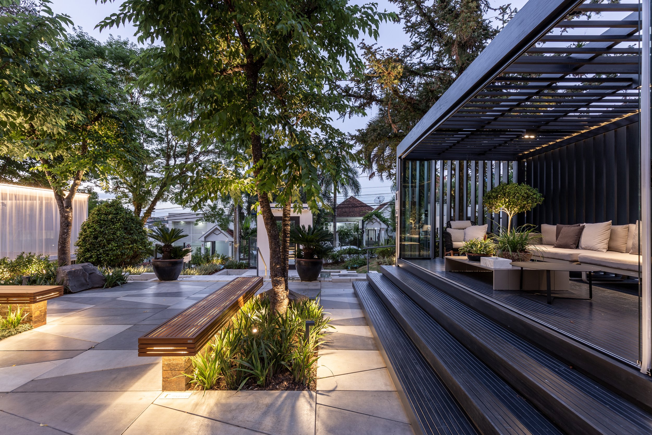 Projeto paisagístico contemporâneo de alto padrão que integra um pavilhão metálico preto com pergolado de lâminas a um amplo jardim de piso em placas de pedra natural cinza, pontuado por canteiros orgânicos com pau-ferro adulto, buxinhos topiados e forrações tropicais iluminados por uplighting e balizadores embutidos. Bancos lineares em madeira cumaru com iluminação LED indireta na base criam áreas de estar contemplativas em meio à vegetação exuberante, enquanto vasos cerâmicos negros de grande , estilo Moderno, Tropical, elementos Bancos de madeira cumaru com iluminação LED embutida na base, Pergolado metálico preto com lâminas horizontais, Deck de madeira composta escura com degraus largos, Piso de placas de pedra natural em tons de cinza com juntas abertas, Vasos cerâmicos pretos de grande porte, Sofás de área externa com almofadas em tons neutros, Iluminação de piso tipo balizador embutido, Iluminação de destaque tipo spot para árvores (uplighting), Cortina luminosa em muro lateral, Esquadrias de vidro com perfil metálico preto, Rocha ornamental natural junto aos bancos, Estrutura de container ou volume metálico preto como pavilhão, plantas Pau-ferro, Buxinho, Moreia, Lírio-da-paz, Clúsia, Agapanto, Formio, Podocarpo topiado, Aspidistra, Palmeira-ráfia, ambiente sofisticado, moderno, relaxante, localização jardim, quintal, entrada, área grande, estruturas pergolado, banco, iluminacao, cores preto, cinza, verde-escuro, bege, marrom, branco, piso pedra, deck, pisantes, privacidade cerca_viva, muro_verde, água nenhum