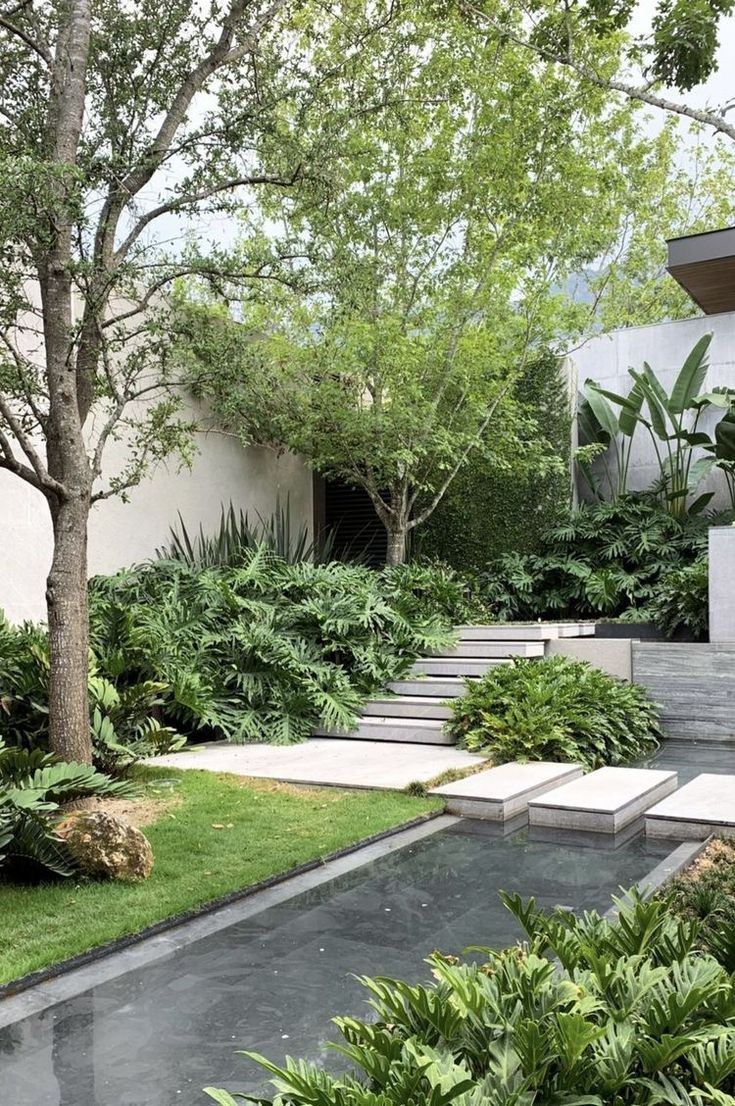 Projeto paisagístico contemporâneo de grande porte que integra arquitetura minimalista com volumetria tropical exuberante, criando uma transição orgânica entre níveis por meio de escadaria em concreto aparente ladeada por maciços densos de filodendros, samambaias e agaves sob a copa de árvore de grande porte. O espelho d'água linear com pisantes flutuantes em concreto claro confere sofisticação e frescor ao conjunto, refletindo a vegetação circundante e conectando visualmente o piso inferior ao , estilo Moderno, Tropical, elementos Escada em degraus de concreto aparente com acabamento liso, Espelho d'água linear com borda em pedra escura, Pisantes retangulares de concreto claro sobre espelho d'água, Canteiros elevados com bordas em concreto, Muro de alvenaria com acabamento em reboco branco, Parede verde vertical com trepadeira, Rocha ornamental natural junto ao gramado, Piso em placas de pedra ardósia escura, Beiral em madeira na edificação ao fundo, plantas Guapuruvu, Filodendro-guaimbê, Bananeira-de-jardim, Samambaia, Agave, Unha-de-gato, Grama-esmeralda, Filodendro-xanadu, Costela-de-adão, ambiente sofisticado, moderno, relaxante, localização jardim, entrada, piscina, área grande, estruturas iluminacao, cores Verde intenso, Verde claro, Cinza escuro, Branco, Bege, Marrom madeira, Verde musgo, piso grama, pisantes, pedra, porcelanato, privacidade muro_verde, cerca_viva, água espelho_dagua