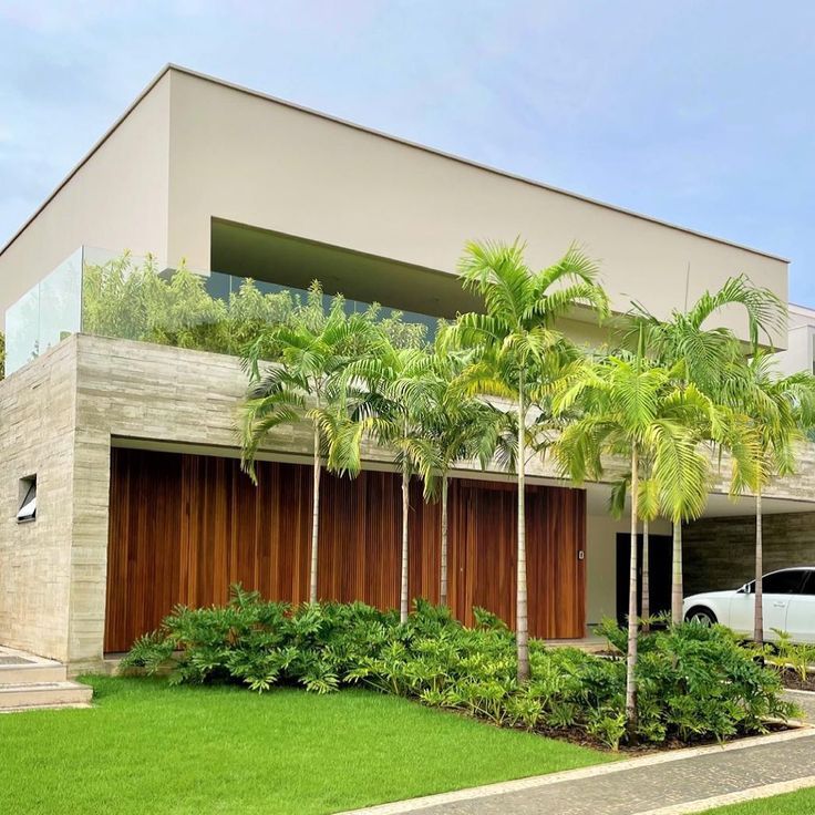 Projeto paisagístico de fachada residencial contemporânea que integra um maciço tropical composto por palmeiras-areca e ráfis enfileiradas à frente de painéis de madeira ripada em cumaru, criando um jogo de texturas verticais sofisticado entre o vegetal e o construtivo. O canteiro frontal é preenchido por forrações densas de filodendros, singônios e clúsias que formam um tapete verde volumoso sob as palmeiras, contrastando com o gramado esmeralda impecavelmente aparado. A varanda superior recebe, estilo Moderno, Tropical, elementos Parede revestida em pedra natural travertino cinza, Painéis de madeira ripada cumaru na fachada, Guarda-corpo em vidro temperado no pavimento superior, Canteiro frontal com forrações tropicais adensadas, Piso intertravado de concreto na área de estacionamento, Volumetria arquitetônica em concreto aparente e massa branca, Vaga de garagem coberta lateral com veículo branco, plantas Palmeira-imperial, Ráfis, Palmeira-areca, Grama-esmeralda, Singônio, Filodendro, Clúsia, Helicônia, Moréia, Pândano, ambiente moderno, sofisticado, localização fachada, jardim, entrada, área grande, estruturas iluminacao, cores Verde intenso, Marrom madeira cumaru, Cinza pedra natural, Branco gelo, Verde claro (grama), Bege areia, piso grama, pedra, privacidade cerca_viva, água nenhum