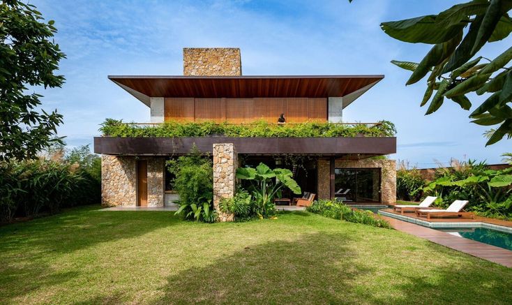 Projeto paisagístico tropical contemporâneo integrado a uma residência de volumetria marcante com laje em balanço, pedra natural e forro de madeira, onde massas de vegetação exuberante — heliconias, bananeiras-ornamentais, filodendros e samambaias — criam transições orgânicas entre a arquitetura brutalista e o jardim. A jardineira suspensa no segundo pavimento transborda com espécies pendentes como jiboias e ripsális, formando uma cortina verde que suaviza a empena de aço corten. O amplo gramado, estilo Moderno, Tropical, elementos Piscina retangular com borda infinita em tom escuro, Espreguiçadeiras de madeira teca com estofado claro, Muro de pedra natural tipo São Tomé ou moledo, Laje em balanço com forro de madeira cumaru, Jardineira suspensa no segundo pavimento com plantas pendentes, Pilares de pedra natural rústica, Esquadrias de madeira de correr no pavimento superior, Chaminé revestida em pedra natural, Amplo gramado plano como tapete verde frontal, plantas Helicônia, Bananeira-ornamental, Palmeira-leque, Costela-de-adão, Filodendro, Clúsia, Singônio, Ripsális, Jiboia, Asplenio, Alpínia, Grama-esmeralda, Pingo-de-ouro, Agapanto, Lírio-da-paz, Taioba-ornamental, Samambaia, Unha-de-gato, Jabuticabeira, Mangueira, ambiente relaxante, moderno, sofisticado, localização jardim, fachada, piscina, varanda, área grande, estruturas iluminacao, cores verde-escuro, verde-claro, bege-pedra, marrom-madeira, cinza-concreto, azul-piscina, branco-areia, ferrugem-corten, piso grama, pedra, privacidade cerca_viva, muro_verde, água piscina