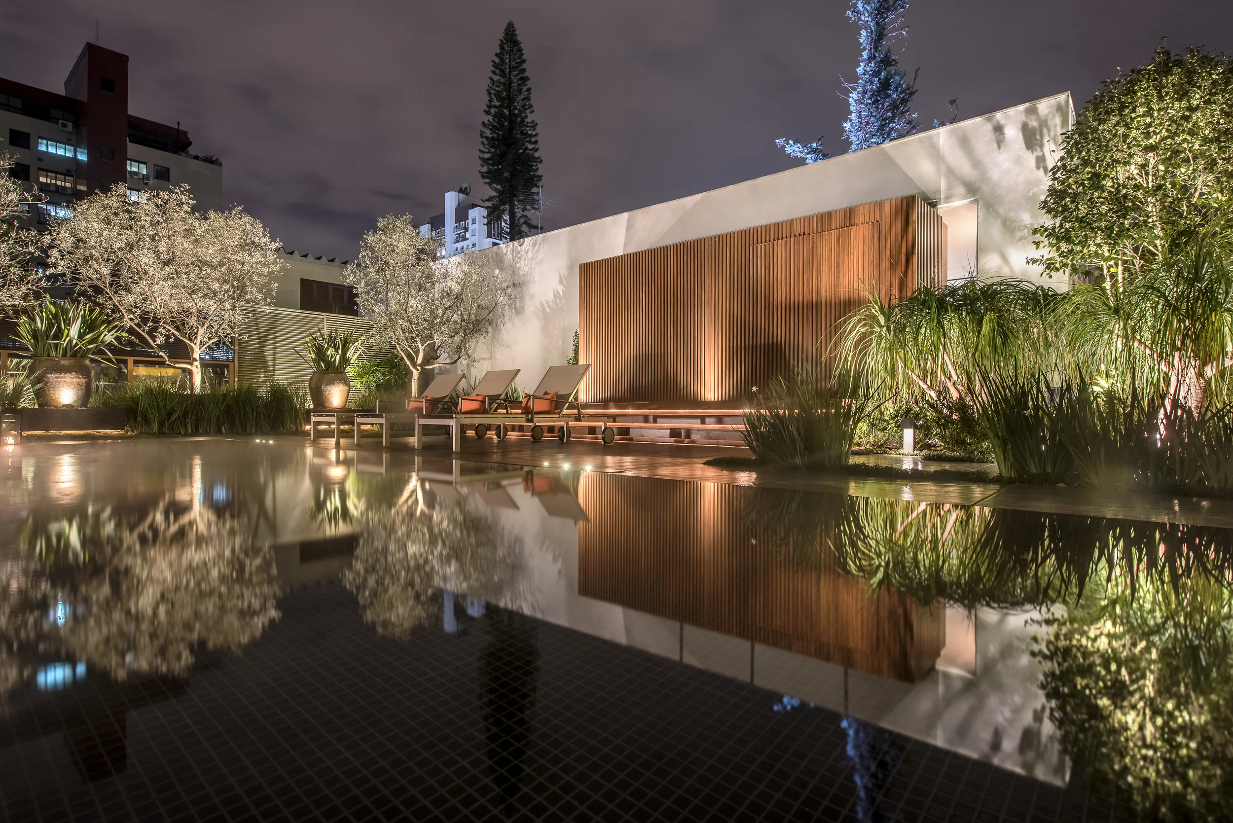Projeto paisagístico contemporâneo de alto padrão com espelho d'água revestido em pastilhas pretas que cria reflexos dramáticos da vegetação e da arquitetura, emoldurado por um imponente painel ripado em madeira cumaru que funciona como elemento focal e integra a área de estar com espreguiçadeiras. A composição vegetal mescla oliveiras adultas com copa escultural, pinheiro-de-norfolk como ponto vertical marcante e maciços de capim-do-texas e papiros que suavizam as bordas do espelho d'água, cria, estilo Moderno, Tropical, elementos Painel ripado em madeira cumaru vertical com iluminação embutida, Muro branco rebocado como pano de fundo, Espreguiçadeiras de madeira com estofado terracota, Espelho d'água com revestimento em pastilha preta, Vasos cerâmicos de grande porte, Iluminação cênica uplighting nas árvores, Balizadores em LED embutidos no piso, Canteiros elevados com bordadura em concreto, Piso em deck de madeira na área de descanso, plantas Oliveira, Pinheiro-de-norfolk, Moreia, Capim-do-texas, Palmeira-ráfis, Dracena, Papiro, Capim-chorão, Agave, Grama-preta, ambiente sofisticado, moderno, relaxante, localização piscina, jardim, muro, área grande, estruturas iluminacao, banco, cores marrom-madeira, branco, verde-escuro, preto, terracota, âmbar, piso deck, porcelanato, pedra, privacidade muro_verde, cerca_viva, água espelho_dagua, piscina